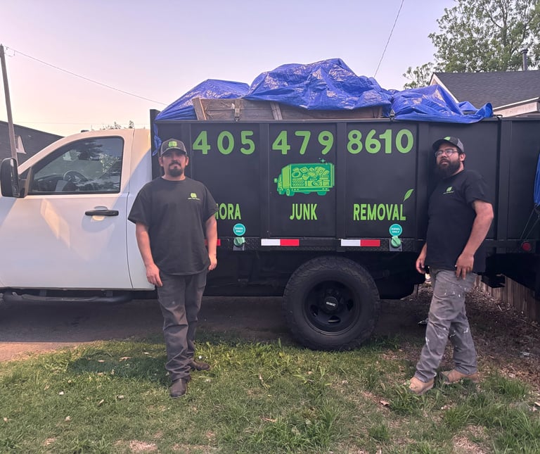 Two workers stand beside a black junk removal truck with green text and a blue tarp-covered load at dusk