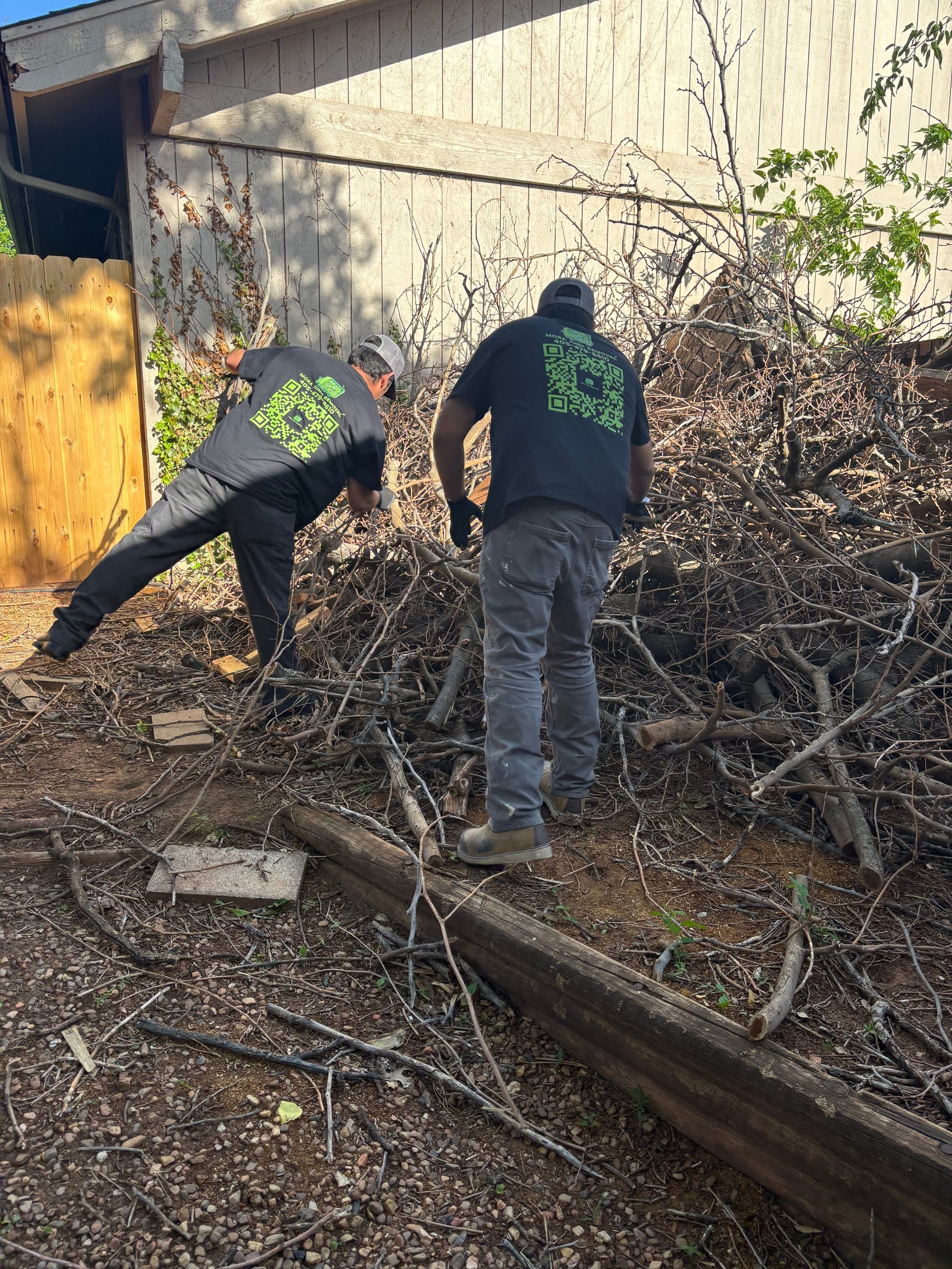 Two volunteers in green-logo black shirts clearing branches and debris from a wooden pile against an industrial building.