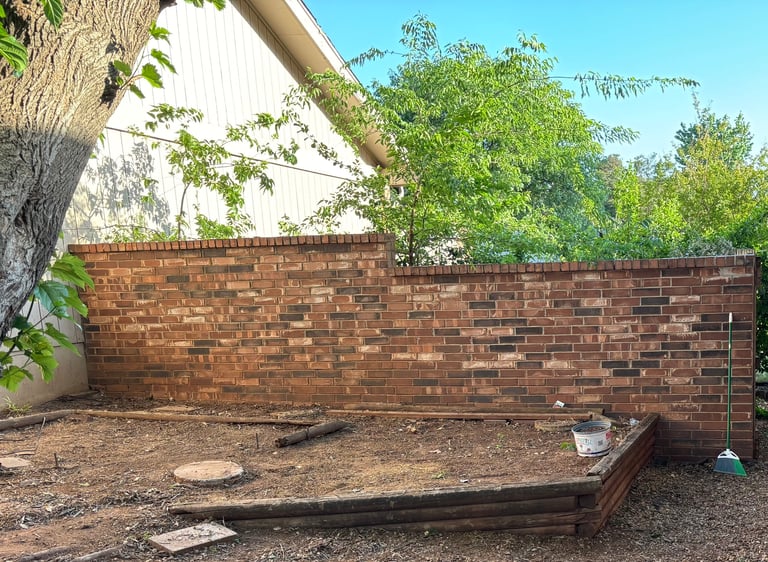 Red brick wall in a backyard with wooden bench, tree, and gravel ground under blue sky