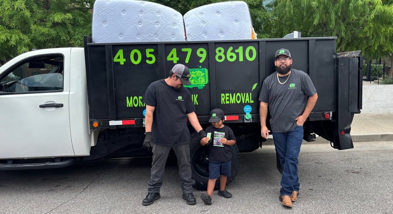 Three workers standing in front of a black junk removal truck with contact number 405 479 8610 displayed in green text